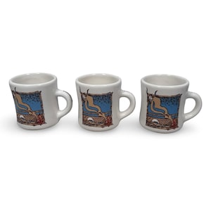 May include: Three white ceramic mugs with a curved handle. Each mug features a rectangular image with a blue sky, a brown and beige landscape, and a tan figure. The mugs are arranged in a row.