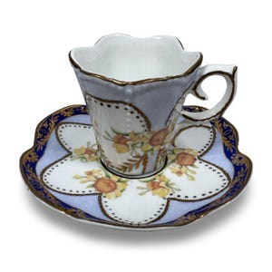 May include: A delicate teacup and saucer set. The cup and saucer feature a light blue and white floral design with gold trim. The cup has a unique scalloped rim and a curved handle. The saucer has a matching floral pattern.