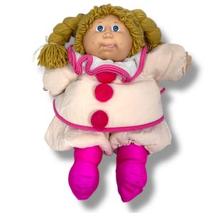 May include: A vintage doll with blonde yarn hair styled in pigtails. The doll has blue eyes and a slightly protruding tongue. It wears a light pink outfit with a white collar, pink trim, and three large pink pom-poms. The doll's legs are covered in bright pink fabric.