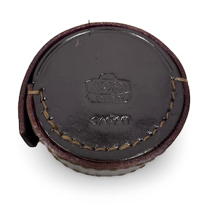 May include: A round, dark brown leather case with a stitched edge. The case has a flap and features embossed text that reads "Nippon Tokyo" and "Japan". The case appears to be for a camera lens.