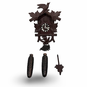 May include: A dark brown wooden cuckoo clock featuring carved leaf motifs, a bird perched on the roof, and a bird at the base. The clock face has white Roman numerals and hands. Two pinecone-shaped weights and a leaf-shaped pendulum are included.