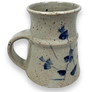 May include: A speckled ceramic mug with a wide handle and a flared rim. The mug is decorated with blue floral designs. The mug has a cylindrical shape with a band around the middle. The mug is a neutral color with dark speckles.