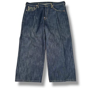 May include: Dark wash denim jeans with a wide leg design. The jeans feature classic five-pocket styling, a button and zipper closure, and light-colored stitching. The denim has a slightly faded appearance, and the jeans are displayed on a white background.