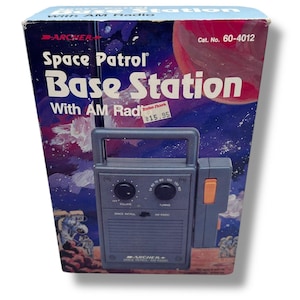 May include: Vintage "Space Patrol Base Station" AM radio in its original box. The box features a space-themed graphic with a planet and astronauts. The radio is gray with a handle and orange accents.