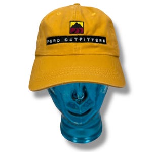 May include: Mustard yellow baseball cap with a curved brim. The cap features a rectangular patch with the words "FORD OUTFITTERS" in white text on a black background. Above the text is a mountain logo with yellow, red, and black colors.