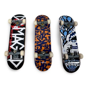 May include: Three finger skateboards with grey metal trucks and white wheels. The left board is black with white text reading DAMAGED. The center board has an orange and blue graffiti pattern. The right board features a blue and white graphic.