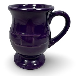 May include: A deep purple ceramic mug with a pedestal base and a curved handle. The mug has a textured design around the middle and a glossy finish. The mug is designed for hot beverages.
