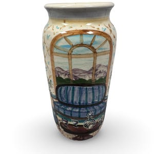 May include: A hand-painted ceramic vase with a detailed interior scene. The design features a window with a mountain view, a blue striped sofa, and a table with glasses. The vase has a light blue rim and a cream-colored background with floral accents.