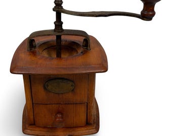 May include: A wooden coffee grinder featuring a square base, a small pull-out drawer with a round knob, and a metal crank handle with a dark wooden knob. A brass-colored oval plate is centered on the front of the wooden body, and the top has a circular opening for beans.