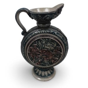 May include: A decorative, round pitcher with a handle and spout. The dark green and silver pitcher features a detailed relief scene of hunters and a boar. The base and spout have ornate designs.