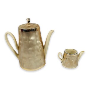May include: A vintage tea set featuring a large teapot and a smaller creamer. The set has a pearlescent finish with silver trim and a cream-colored handle and spout. The teapot has a lid with a decorative finial.