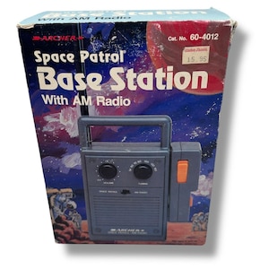 May include: Vintage Space Patrol Base Station AM radio in its original box. The box features a space-themed illustration with a red planet and astronauts. The radio is gray with a handle, volume and tuning knobs, and an orange button.
