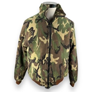 May include: Camouflage-patterned jacket with a full-length zipper. The jacket features a high collar and brown accents on the cuffs and hem. The camouflage design includes shades of green, brown, and tan.