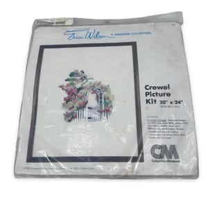 May include: A sealed crewel picture kit by Erica Wilson, featuring a framed design of a summer cottage scene with a white picket fence and floral archway. The kit measures 20 x 24 inches.