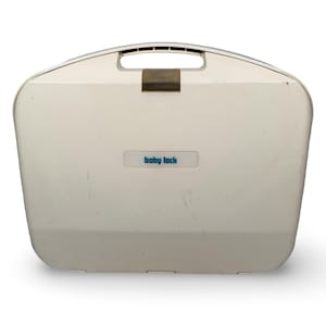 May include: A white, hard-shell carrying case with a top handle and a central latch. The front features a rectangular label with the text 'baby lock' printed in blue. The case has a smooth, slightly curved rectangular shape with minor surface wear.