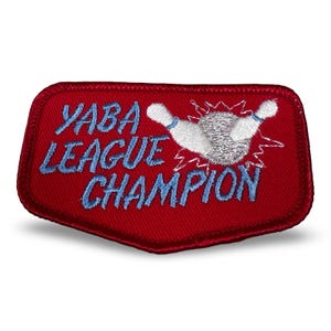 May include: A red embroidered patch with the text "YABA LEAGUE CHAMPION" in blue script. The patch features a bowling pin and ball design in white and silver. The patch has a unique shape with a red border.