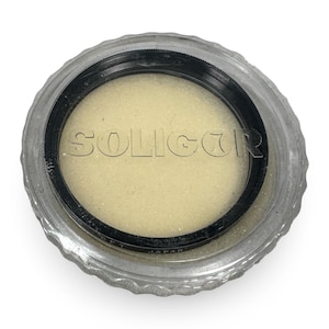May include: A SOLIGOR camera lens filter in a clear plastic case. The filter is black and the case is clear with a ridged edge. The word SOLIGOR is embossed on the filter. The filter is surrounded by a light yellow foam.