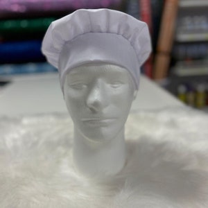 May include: A white chef's hat with a rounded crown and a pleated brim. The hat is made of a smooth, opaque fabric and is displayed on a white mannequin head. The background is blurred, with hints of colorful fabric and other items.