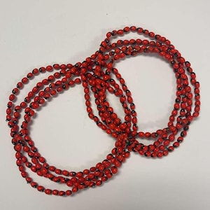 Puede incluir: Varias hebras de collares de cuentas de semillas rojas y negras se disponen sobre una superficie blanca. Las cuentas, de forma ovalada, tienen un exterior rojo vibrante y una mancha negra en un extremo. Los collares están entrelazados, creando un efecto de capas.