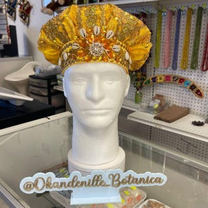 May include: A yellow head covering with gold sequins, shells, and a central jewel-like embellishment. The headpiece is displayed on a white mannequin head. The sign in front reads "@Okandeville_Botanica".