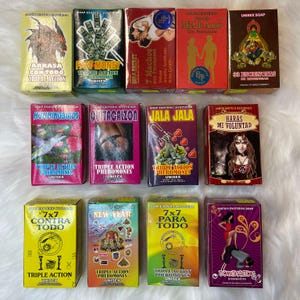 May include: Twelve small, colourful boxes of soap with various designs and text. The boxes are arranged in three rows of four. The text on the boxes includes "Arrasa con Todo", "Fast Money", "7 Machos", "Miel de Amor", "33 Esencias", "Hummingbirds", "Quitacalzon", "Jala Jala", "Haras Mi Voluntad", "7x7 Contra Todo", "New Year", "7x7 Para Todo", and "Domination".