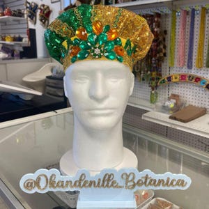 Pode incluir: Um chapéu decorativo com lantejoulas verdes e renda dourada, adornado com pedras preciosas laranja e verdes. O chapéu é exibido em uma cabeça de manequim branca. A placa abaixo diz "@Okandeville_Botanica".