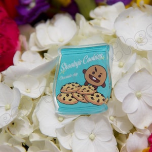 May include: A light blue sticker featuring a cartoon cookie bag with the text "Shooky's Cookies" and a cartoon character. The sticker also shows chocolate chip cookies. The sticker is set against a backdrop of white and pink flowers.