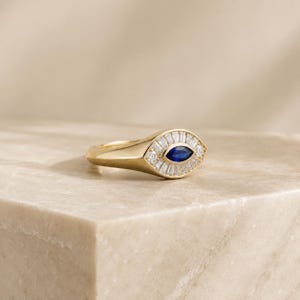 May include: A gold ring featuring a central marquise-cut blue sapphire surrounded by a halo of baguette-cut white diamonds. The ring has a polished gold band and a tapered setting that frames the stones in an eye-shaped design with clear detail.
