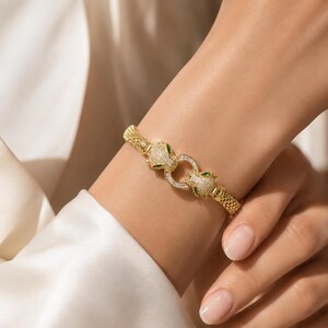 May include: A gold-toned bracelet featuring a flexible mesh band and a central clasp designed as two facing panther heads. The panther heads are encrusted with small clear stones and feature green stone accents for the eyes, connected by a ring.