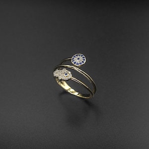 May include: A gold ring with two evil eye charms. One charm is a circle with a blue eye and the other is a hand with a blue eye. The ring is set with clear stones.
