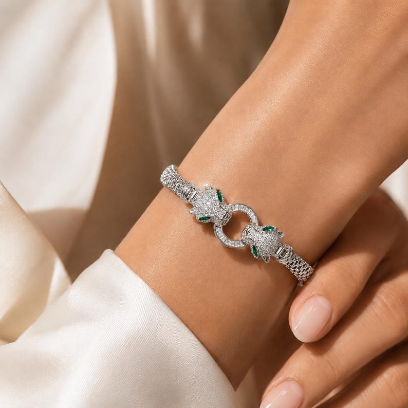 May include: A silver-toned bracelet featuring a mesh band and two panther head motifs encrusted with clear stones and accented with green stones for the eyes. The two heads meet at a central circular clasp adorned with baguette-cut clear stones.