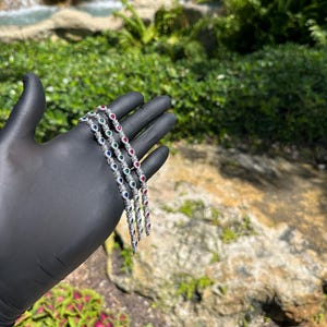 May include: Four silver bracelets with alternating red, green, blue, and clear gemstones. The bracelets are held in a black gloved hand against a backdrop of green foliage and a small waterfall.