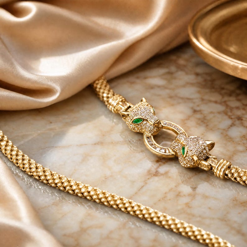 May include: A gold-toned chain necklace featuring a central clasp designed as two facing panther heads. The panther heads are encrusted with small clear stones and feature vibrant green stone eyes, connected by a circular link with baguette stones.