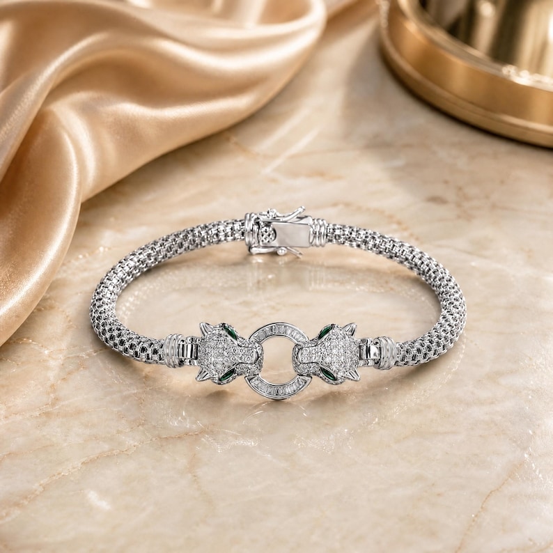 May include: A silver-toned mesh bracelet featuring two panther head motifs encrusted with clear stones and small green accents. The panther heads are connected by a central circular ring, and the bracelet includes a secure metal box clasp closure.