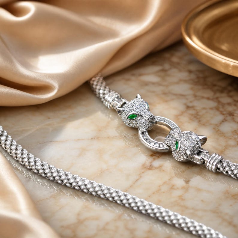 May include: A silver-toned mesh chain necklace featuring a clasp designed as two panther heads. The panther heads are encrusted with small clear stones and feature vibrant green marquise-cut stones for the eyes, connected by a circular ring.
