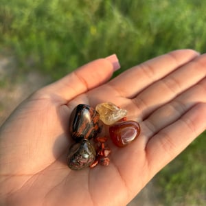 May include: A collection of tumbled stones in various colors, including red, brown, black, and yellow. The stones are arranged in a hand, with the largest stone in the center.