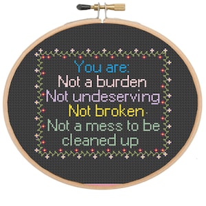 Op de afbeelding: Een zwarte borduurring met een witte achtergrond en kleurrijke tekst die "You are: Not a burden Not undeserving. Not broken. Not a mess to be cleaned up." luidt.