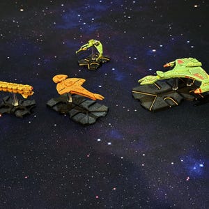 May include: Four miniature plastic starships mounted on black hexagonal bases. The collection includes a long yellow freighter, an orange vessel with a flat hull, a small green scout ship, and a large green bird-of-prey style starship.