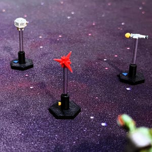 May include: Three miniature game pieces mounted on silver rods and black hexagonal bases. One piece is a white spherical structure, one is a red star-shaped object, and one is a white cylindrical shape with a yellow tip, all on a dark purple surface.