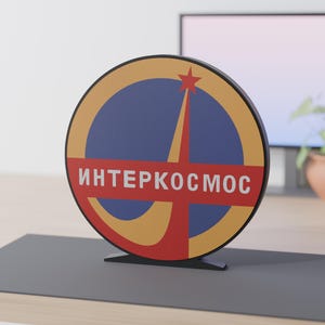 May include: A circular sign featuring the Interkosmos logo with a blue background, a yellow crescent shape, and a red star. A horizontal red bar across the center displays the white Cyrillic text "ИНТЕРКОСМОС". The sign is mounted on a black base.