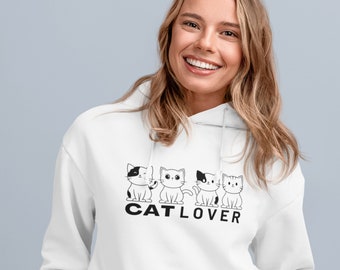 以下が含まれることがあります： 白いフード付きスウェットシャツ。黒と白の漫画の猫が4匹と、下に「CAT LOVER」の文字がプリントされています。