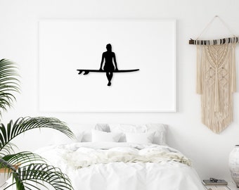 May include: Black silhouette of a person sitting on a surfboard, framed and hanging on a white wall above a white bed with white bedding.