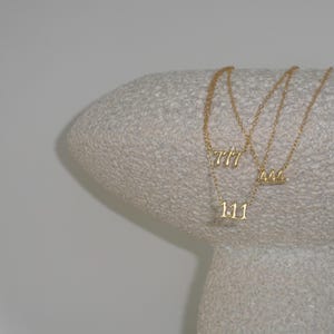 May include: Three gold necklaces with number pendants: 111, 444, and 777. The necklaces are displayed on a textured, light-coloured surface. The pendants are delicate and feature small embellishments.