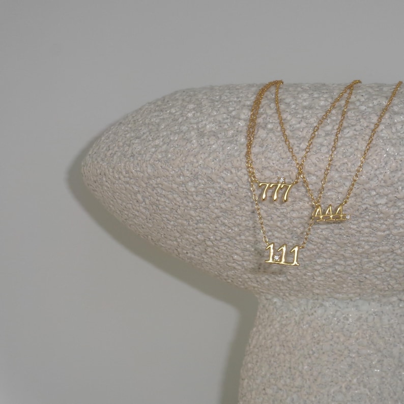 May include: Three gold necklaces with number pendants: 111, 444, and 777. The necklaces are displayed on a textured, light-coloured surface. The pendants are delicate and feature small embellishments.