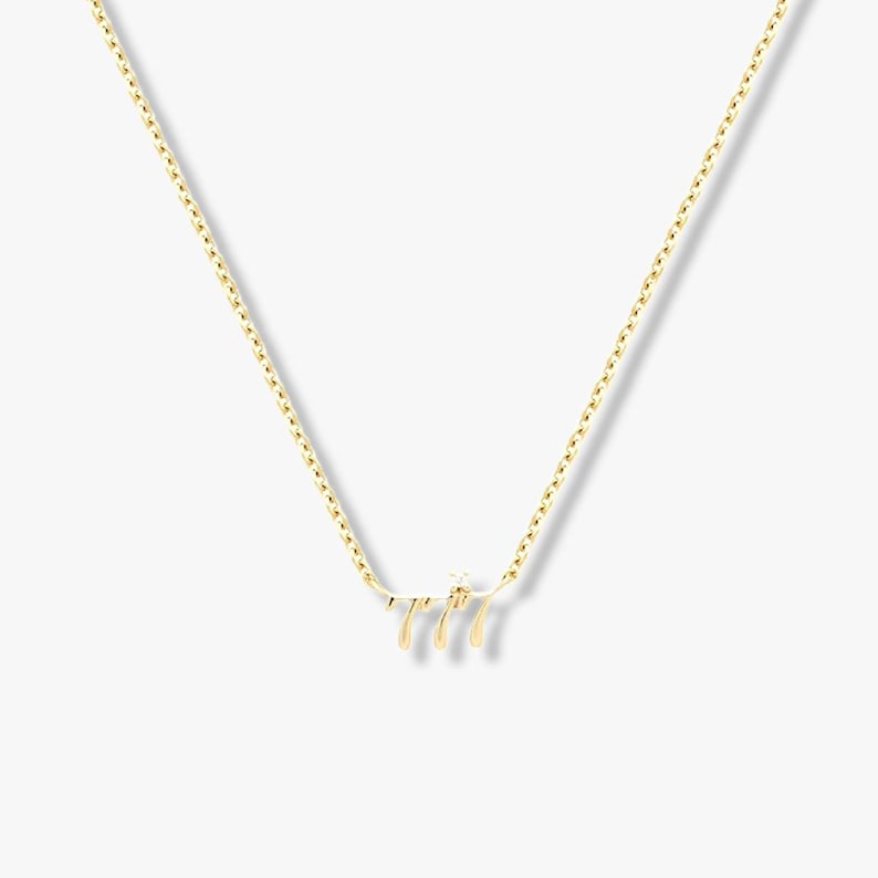 May include: A gold-coloured necklace with a pendant displaying the number "777" in a decorative style. The necklace has a fine chain and is set against a plain backdrop. The pendant includes a small, sparkling detail.