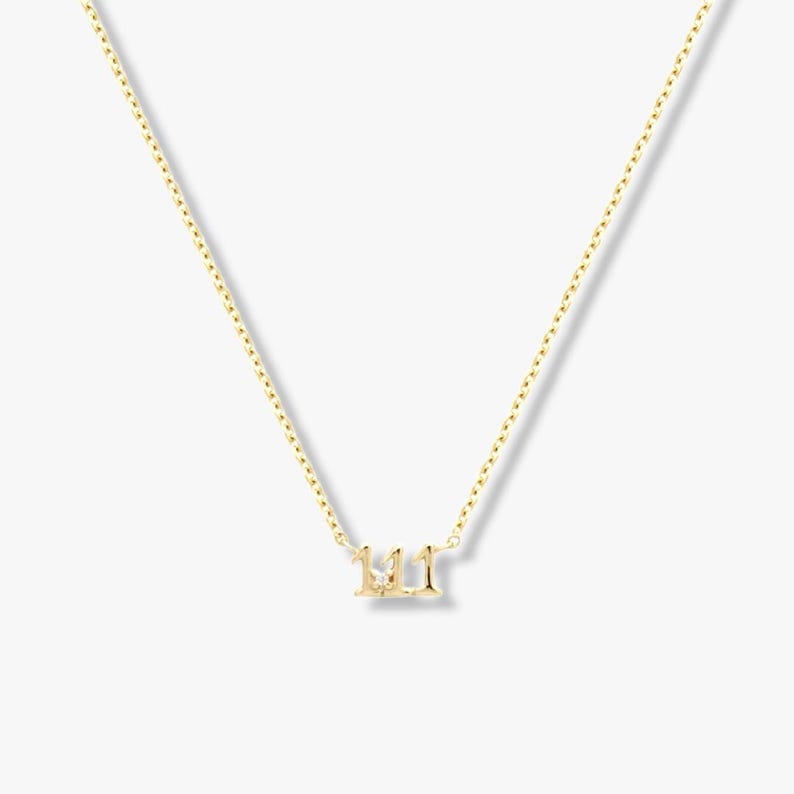 May include: A gold-coloured necklace with a pendant displaying the number "111". The pendant is small and hangs from a fine chain. The necklace is shown against a plain background.