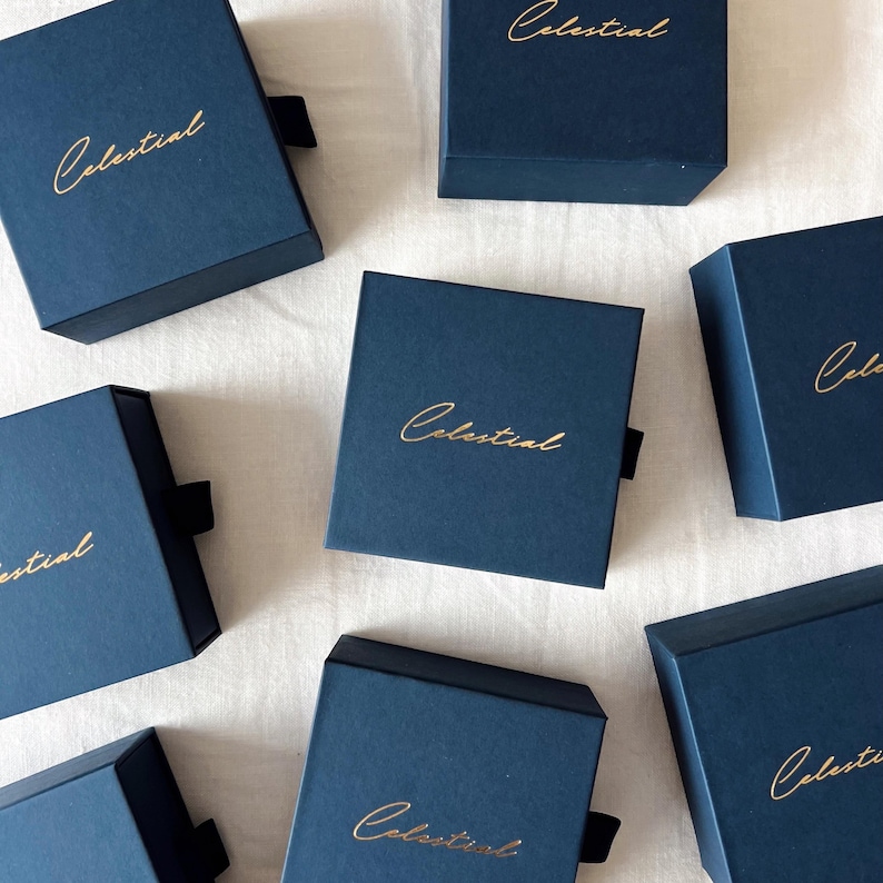May include: Several navy blue square boxes with the word "Celestial" in gold lettering. Each box has a black ribbon pull. The boxes are displayed on a white surface.