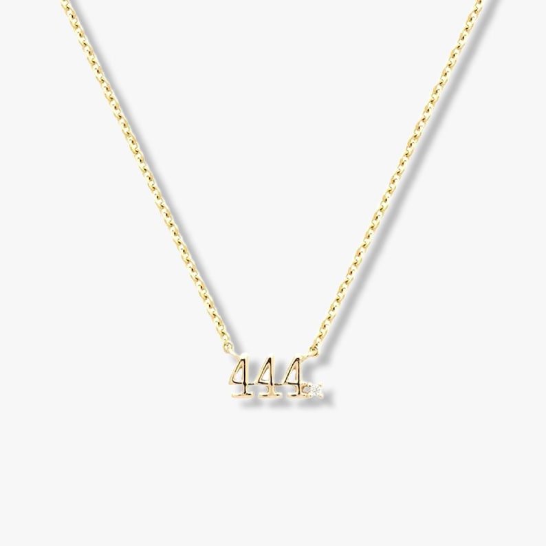 May include: A gold-tone necklace with the number "444" as a pendant, accompanied by a small diamond. The chain is fine and elegant. The necklace is presented on a neutral background. The necklace is suitable for everyday wear.