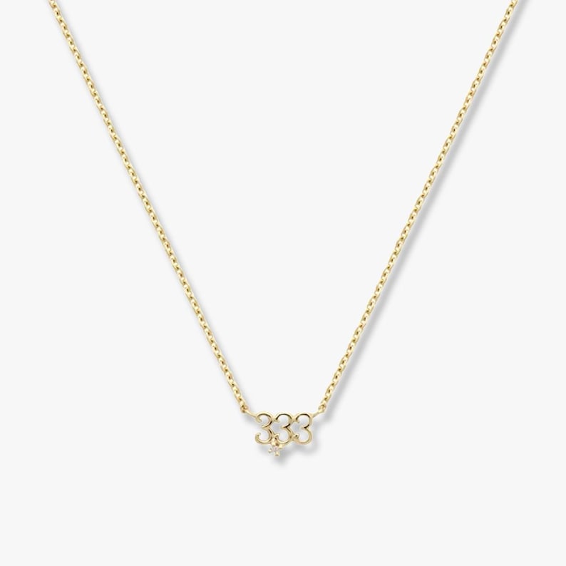 May include: A gold-coloured necklace with a fine chain and a pendant. The pendant is in the shape of the number "333" with a small, sparkling accent. The necklace is shown against a plain background.