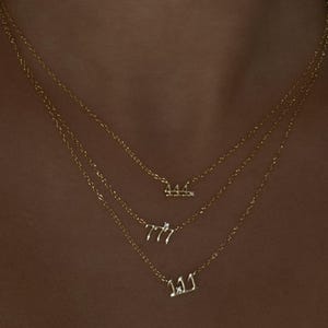 May include: Three gold necklaces with pendant numbers 111, 777, and 444. Each necklace has a fine chain. The pendants are in a script font and have small, clear gemstones. The necklaces are layered and worn on a person.